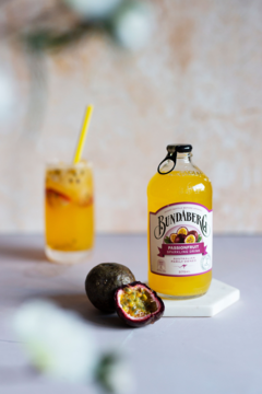 Butelka Bundaberg Passionfruit Brew, żółty napój, obok szklanka z kruszonym lodem i świeże passiflory.