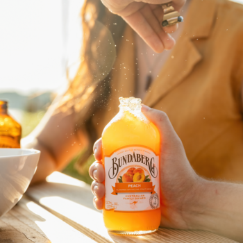 Zbliżenie na butelkę Bundaberg Peach z pomarańczowym napojem, trzymaną w dłoni, z osobą sypiącą cukier.