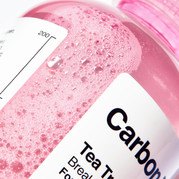 Zbliżenie butelki Carbon Theory Tea Tree Tonic, różowy spieniony płyn i fragmenty białych etykiet.