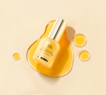 Dwuwarstwowe serum d'Alba White Truffle Double Layer, złota butelka leżąca w kałuży olejku, beżowe tło.