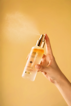 Ręka rozpyla dwufazowe serum d’Alba White Truffle First Spray Serum, z widoczną mgiełką, na żółtym tle.