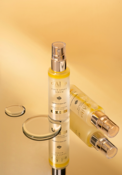 Buteleczka d'Alba White Truffle First Spray Serum, rozświetlające, z żółtym płynem, stoi na błyszczącej złotej powierzchni z kroplami.