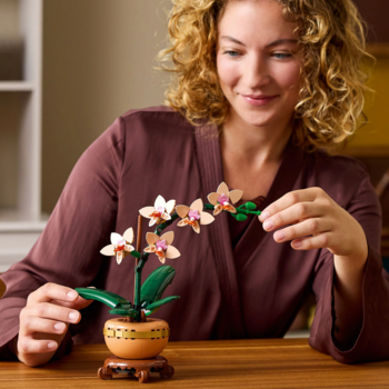 Uśmiechnięta kobieta w brązowej szacie podziwia model orchidei LEGO z różowymi kwiatami w ozdobnej doniczce.