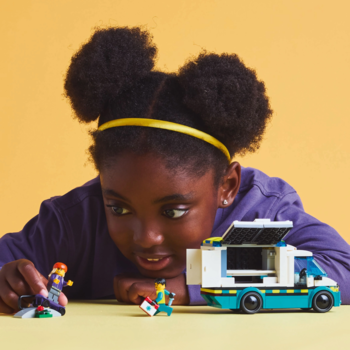 Dziewczynka bawi się zestawem Lego: turkusowy mobilny pojazd z otwartym bokiem i figurki, na żółtym tle.