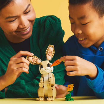 Kobieta i chłopiec budują beżowego LEGO królika z ruchomymi uszami, podając mu marchewkę. Żółte tło.