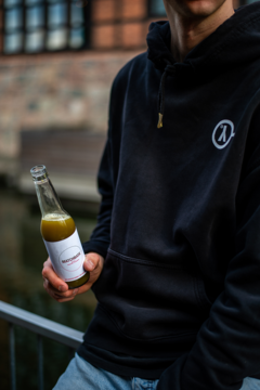 Zielony sok w szklanej butelce z białą etykietą, trzymany przez osobę w czarnej bluzie z kapturem.