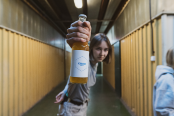 Szklana butelka bursztynowego napoju bezglutenowego z białą etykietą, trzymana przez osobę w industrialnym korytarzu.