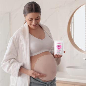 Uśmiechnięta ciężarna kobieta w białym szlafroku trzyma naturalną maskę na brzuch i gładzi swój brzuszek.