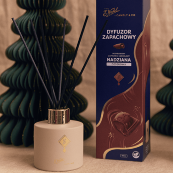 Dyfuzor E. Wedel x Candly & Co Nadziana deserowa, beżowy flakon z patyczkami, obok pudełko, na tle zielonych choinek.