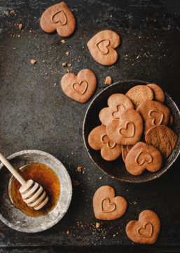 Ciastka korzenne serduszka z miodem, w misce i rozłożone na ciemnym tle, obok miód w naczyniu. Widok z góry.