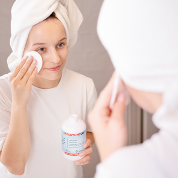 Kobieta używa oczyszczającego toniku Argentum200 z cytrynianem srebra w białej butelce; widok w lustrze.