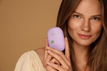 Uśmiechnięta kobieta prezentuje fioletowy krem do rąk handy lab. Energy & Fresh Patchouli Mood, nawilżający, na beżowym tle.