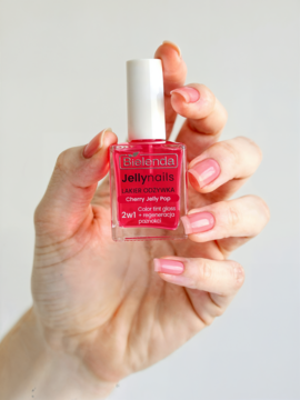 Dłoń trzyma różowy lakier odżywczy Bielenda Jellynails Cherry Jelly Pop w szklanej buteleczce. Różowe paznokcie.