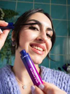 Uśmiechnięta kobieta nakłada pogrubiający tusz LUMENE Wild Forces na rzęsy; zbliżenie na fioletowe opakowanie i aplikację.