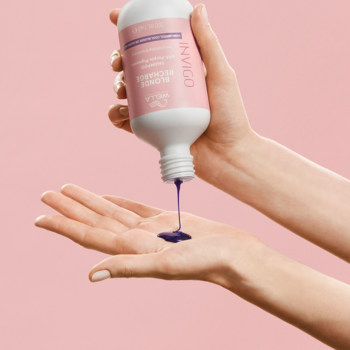 Kobieca dłoń wyciska fioletowy szampon Wella Invigo Blonde Recharge do blond włosów z butelki, różowe tło.