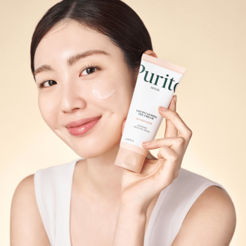 Kobieta prezentuje Purito Daily Go-To Sunscreen, żel-krem z filtrem SPF 50+. Trochę produktu na policzku.