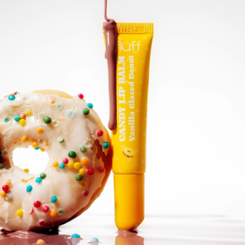 Fluff Candy Lip Balm Vanilla Glazed Donut w żółtej tubce, obok oblany lukrem pączek z posypką na białym tle.