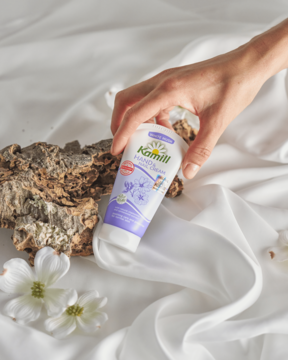 Krem Kamill do rąk z rumiankiem i sandałowcem w białej tubie, trzymany przez dłoń na tle białej tkaniny.