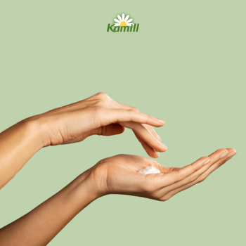 Kamill krem do rąk brzoskwiniowy. Dłonie aplikują biały krem na jasnym zielonym tle, z logo Kamill u góry.
