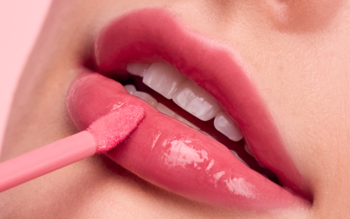 Zbliżenie na usta, różowy błyszczący balsam do ust nakładany aplikatorem na dolną wargę, żelowe wykończenie.