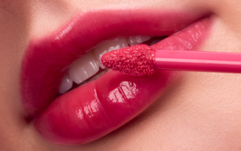 Zbliżenie na lśniące, różowe usta, na które aplikator nakłada błyszczący balsam do ust z efektem pełniejszych warg.