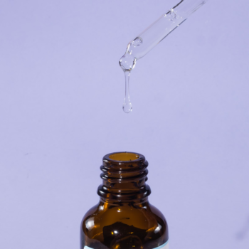 Kropla wodnego serum z azeloglicyną spada z pipety do bursztynowej butelki na fioletowym tle. Rozjaśnia cerę.