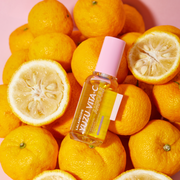 Yuzu Vita-C Ampoule do twarzy, szklana butelka z żółtym płynem i różową nakrętką, otoczona świeżymi owocami yuzu.