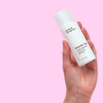 Biała butelka Geek & Gorgeous Smooth Out 12% AHA w dłoni, różowe etykiety, na jasnoróżowym tle.