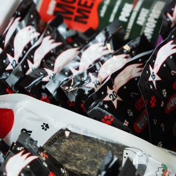 Zbliżenie na kalendarz adwentowy dla psa, stos czarnych saszetek z numerami i motywem komety, obok biała torebka z przysmakiem bez zbóż.