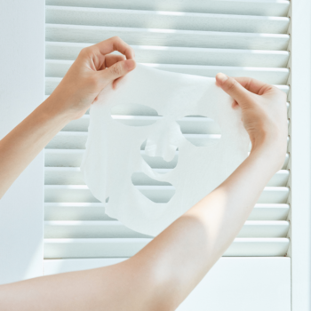 Ręce trzymają rozświetlającą maskę w płachcie Skin1004 Madagascar Centella Tone Brightening Glow Mask przed białymi żaluzjami.