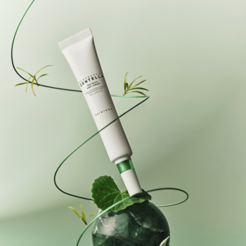 Skin1004 Tea-Trica Spot Cream, biała tubka na niedoskonałości, leży skośnie na zielonym kamieniu, z pnączami.