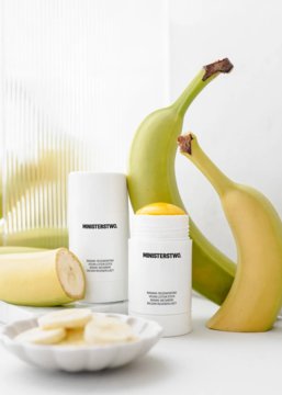 Balsam Ministerstwo Banan w białym sztyfcie, otwarty z żółtym wkładem, na stole z zielonymi bananami.