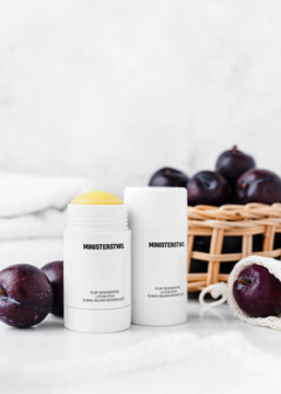 Regenerujący balsam Ministerstwo Śliwka w białym sztyfcie, jeden otwarty, obok świeże śliwki i koszyk.