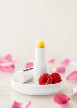 Zbliżenie: żółta pomadka ochronna Ministerstwo Dobrego Mydła Róża-Malina w białym etui, z malinami i różami.