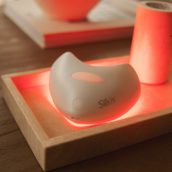 Silk'n LumiLips, ergonomiczne szare urządzenie LED do ust z pulsującym czerwonym światłem na drewnianej tacy.