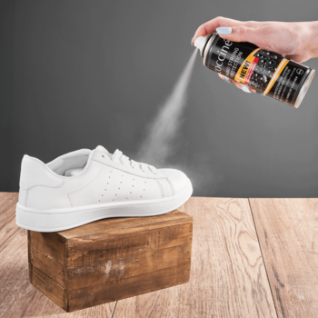 Ręka spryskuje biały but sneakers impregnatem w sprayu na drewnianym blacie, zapewniając wodoodporność.