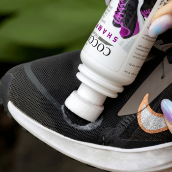 Zbliżenie na dłoń aplikującą szampon Coco Shame do czarnego sneakersa, tworząc pianę. Czyści buty.