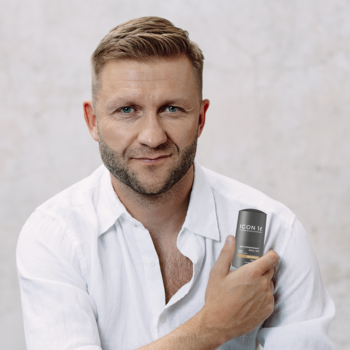 Mężczyzna z brodą w białej koszuli trzyma ciemny produkt do pielęgnacji, patrzy wprost.