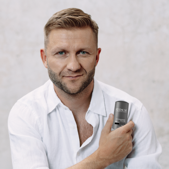Mężczyzna z brodą w białej koszuli trzymający ciemny dezodorant w sztyfcie, patrzący wprost na jasnym tle.