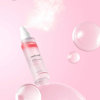 Przezroczysta butelka mediCoup RED B CAPSULE serum mist z różowym płynem, rozpylająca mgiełkę, na różowym tle z bąbelkami.