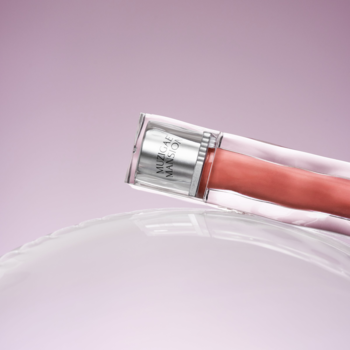 Błyszczyk Muzigae Mansion Icy Glow Noble, brzoskwiniowy w przeźroczystej tubce, na jasnym tle, pod kątem.