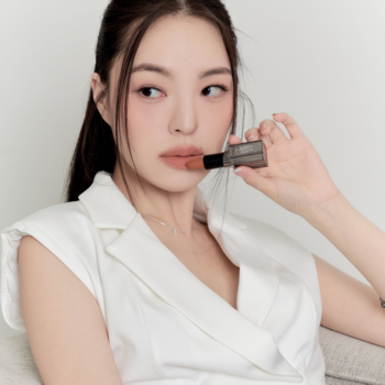 Kobieta prezentuje matową pomadkę Muzigae Mansion Nuddy w szarej obudowie, trzymając ją przy ustach, jasne tło.