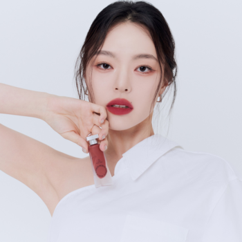 Kobieta z matowymi czerwonymi ustami prezentuje matowy tint do ust Muzigae Mansion Spicy w brązowo-czerwonym opakowaniu.