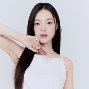 Ujęcie kobiety z długimi włosami, trzymającej matowy tint do ust Muzigae Mansion Posh w przezroczystym opakowaniu z różową zakrętką.