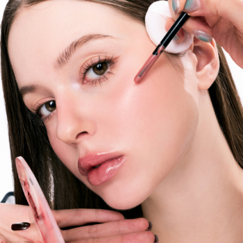 Zbliżenie na modelkę, makijażystka aplikuje płynny róż pędzelkiem na policzek, usta błyszczą.