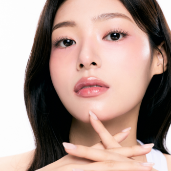 Portret kobiety z delikatnym makijażem, różowym rumieńcem i błyszczącymi ustami, dłonie skrzyżowane na białym tle.