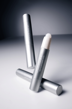 Trzy srebrne sztyfty rozświetlacza Prism Stick. Jeden otwarty, ukazujący biały, precyzyjny aplikator, na jasnoszarym tle.