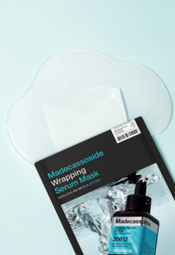Maska w płachcie Madecassoside Wrapping Serum w czarnym opakowaniu i rozłożona, przezroczysta maska cloud, na tle błękitu.