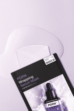 Ciemna maska w płachcie PDRN Wrapping Serum Mask z białym płynem i arkuszem na liliowym tle, widok z góry.