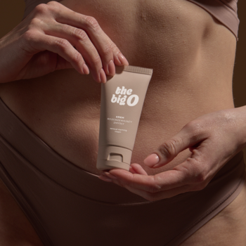 Krem The Big O Daily Sensitivity Enhancing Cream w beżowej tubce, trzymany przez dłonie przy nagim brzuchu.